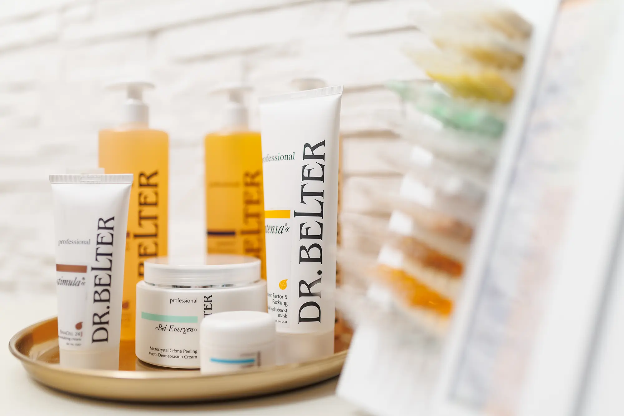 Pappauftsteller mit verschiedenen Ampullen und goldenes Tablett mit Tuben und Spendern der Kosmetikmarke Dr. Belter Cosmetic. Angerichtet sind die beiden Elemente auf einem Schrank vor einer weißen Ziegelmauer.