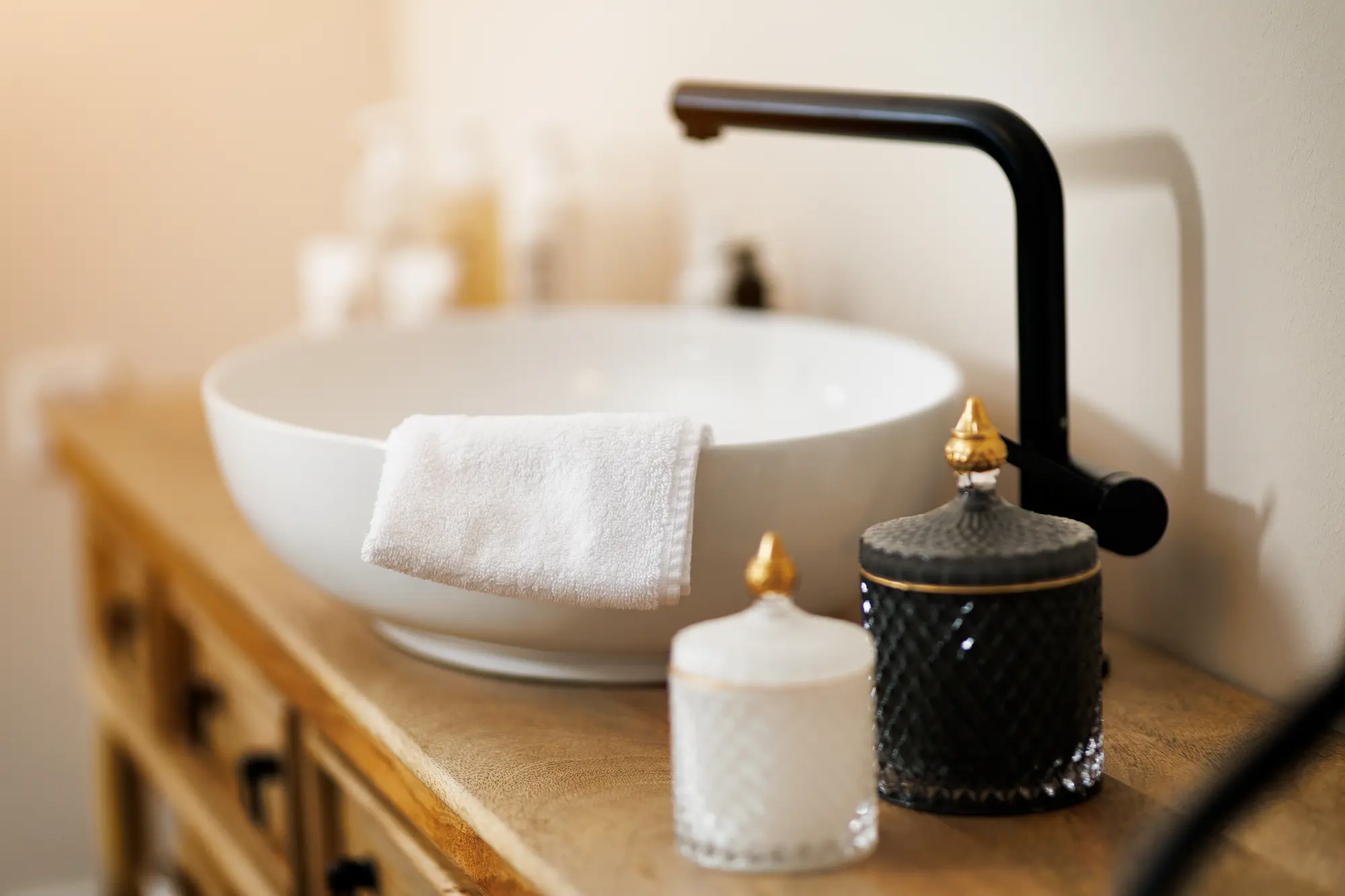 Dunkelbrauner Holzschrank, auf dem sich ein weißes Waschbecken mit einer schwarzen Armatur und ein weißes Handtuch, sowie ein schwarzes und ein weißes Gefäß mit goldenen Griffen befinden