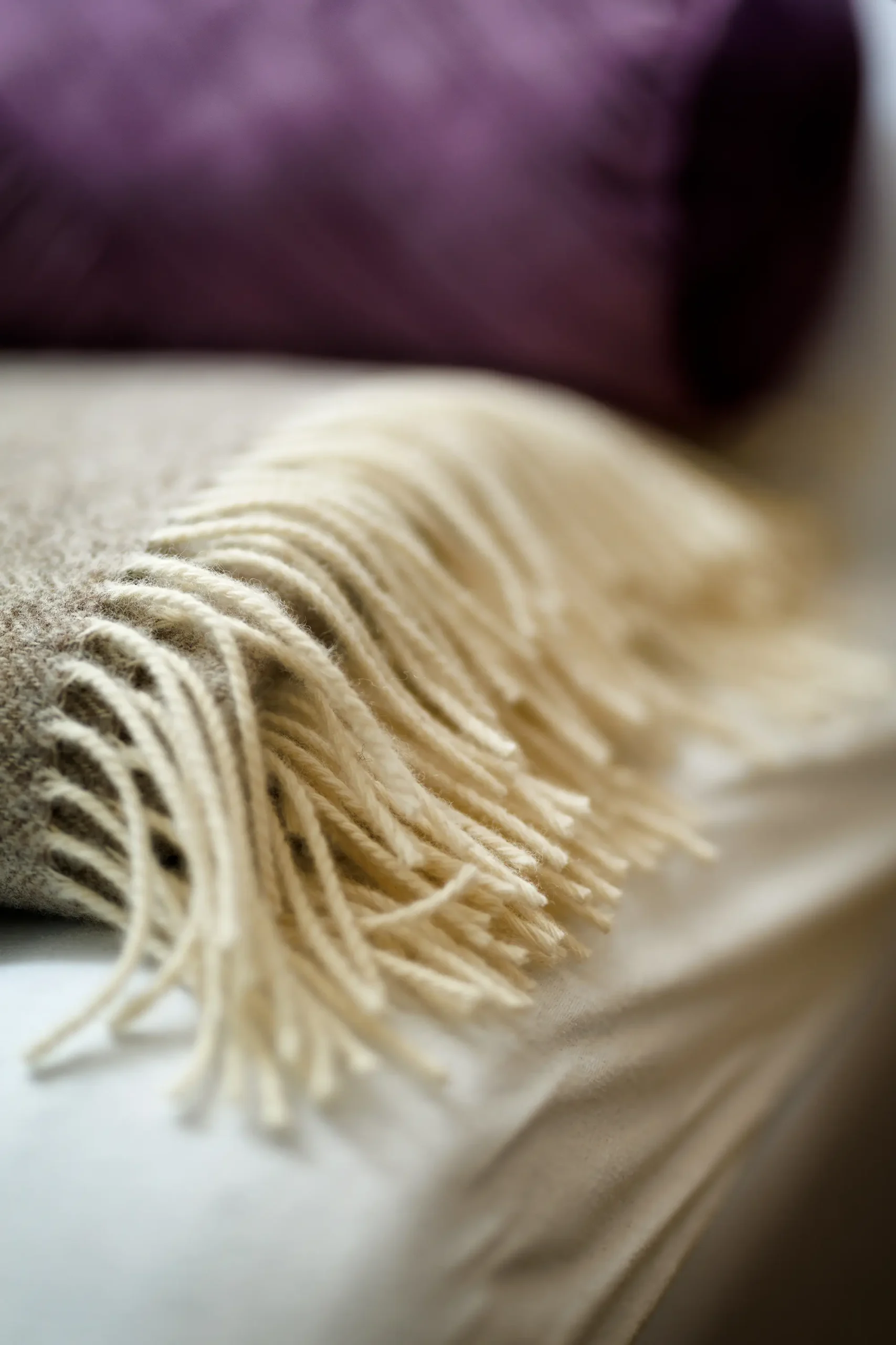 Braune Decke mit weißen Fransen im Vordergrund mit einem lila farbenen Kissen im Hintergrund