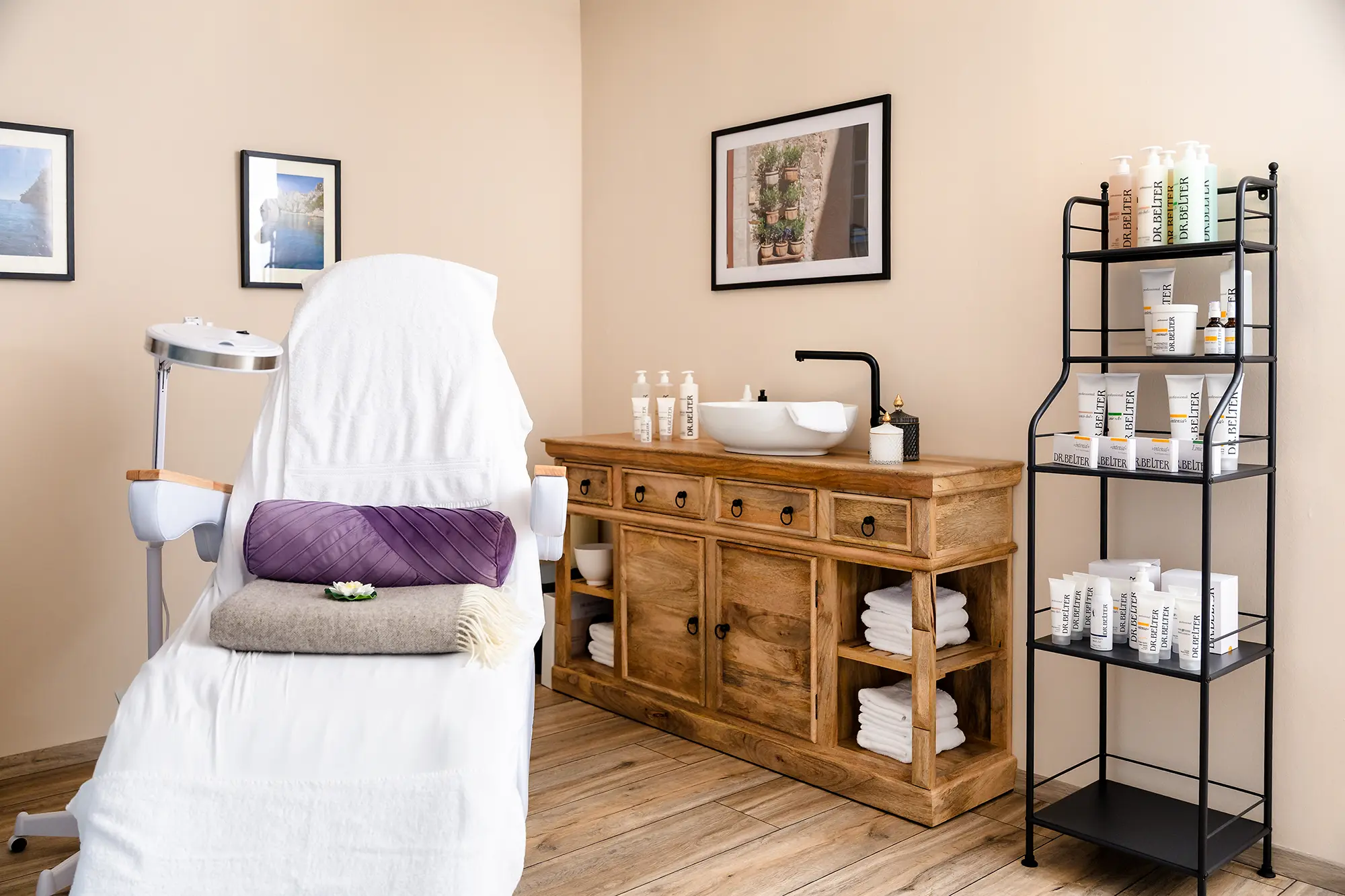 Im Vordergrund eine weiße Liege mit einer Lampe, einem lila farbenen Kissen, einer braunen Decke und einer weißen Lotusblüte. Im Hintergrund ein dunkelbrauner Schrank mit weißem Waschbecken und schwarzer Armatur, sowie Handtücher, Kosmetikprodukten und Gefäßen. Daneben ein schwarzes Metallregal gefüllt mit Kosmetikprodukten der Marke Dr. Belter. An der beigen Wand dahinter befinden sich drei eingerahmte Fotografien.