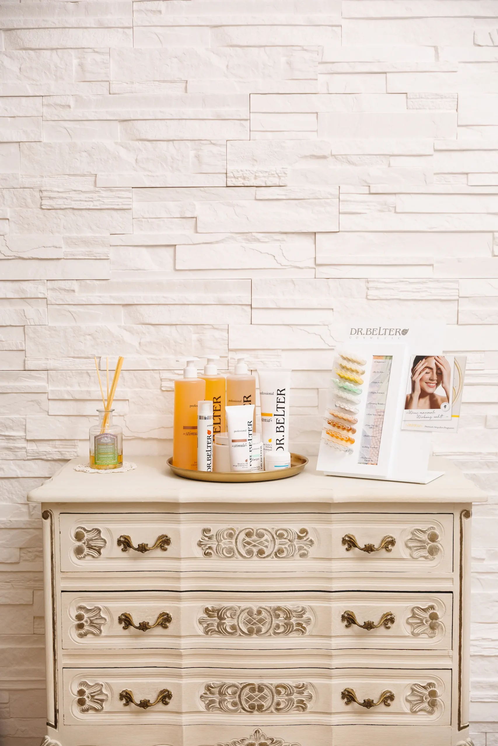 Weiße Wand in Ziegeloptik und weißer Schrank in antiker Optik, auf dem Kosmetikprodukte und Duftstäbchen stehen
