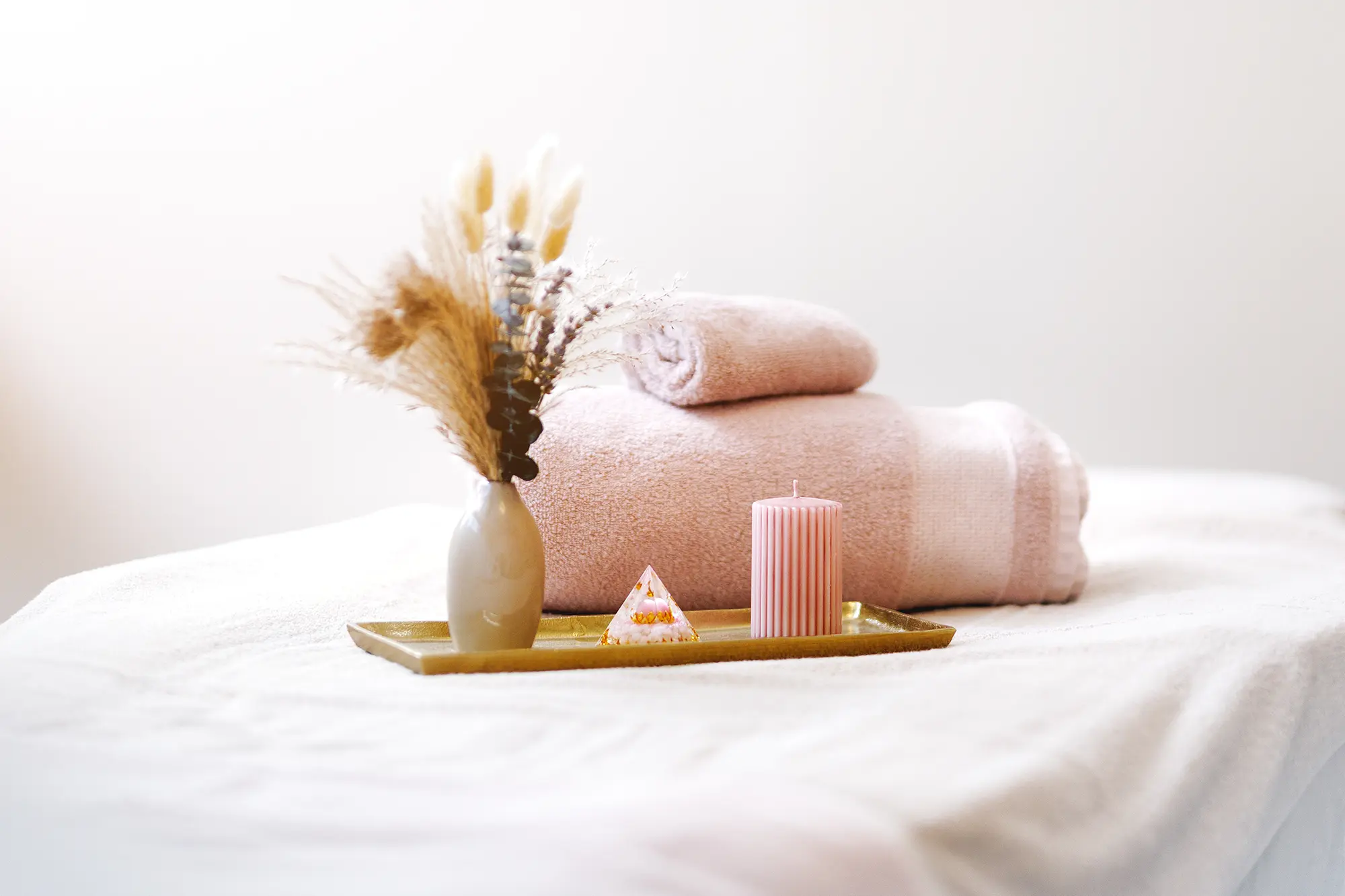 Weißer Hintergrund mit weißer Liege im Vordergrund, auf der sich zwei rosa farbene Handtücher und ein goldenes Tablett mit einer rosa farbenen Kerze, einer kleinen Dekopyramide aus Glas und eine beige Vase mit Blumen befinden