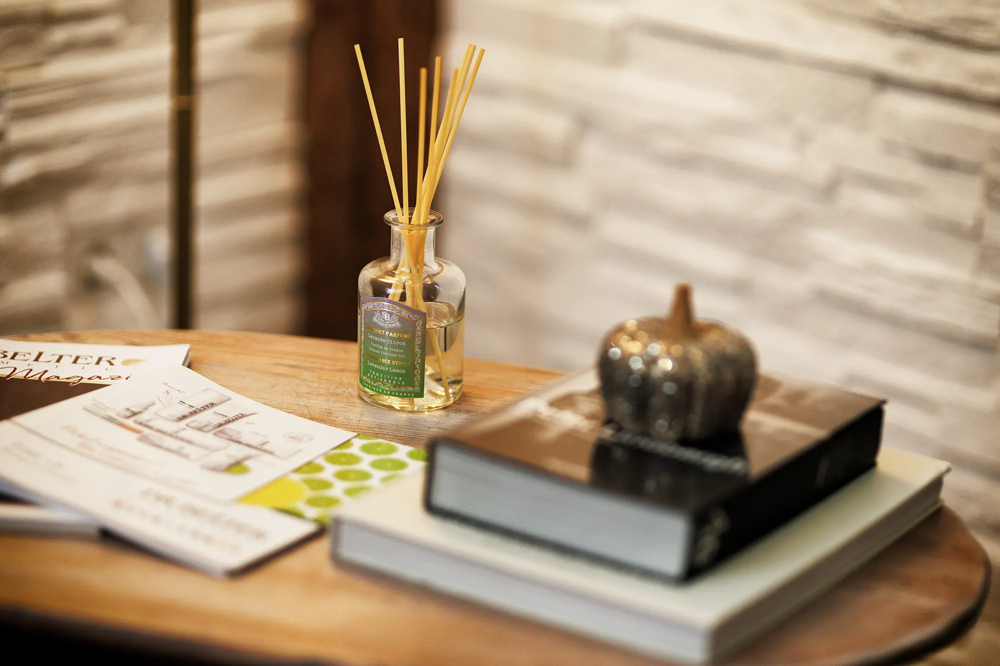 Dunkelbrauner Beistelltisch aus Holz auf dem sich Duftstäbchen, zwei Bücher, ein kleiner schwarzer Kürbis als Dekoration und Magazine der Kosmetikmarke Dr. Belter Cosmetic befinden