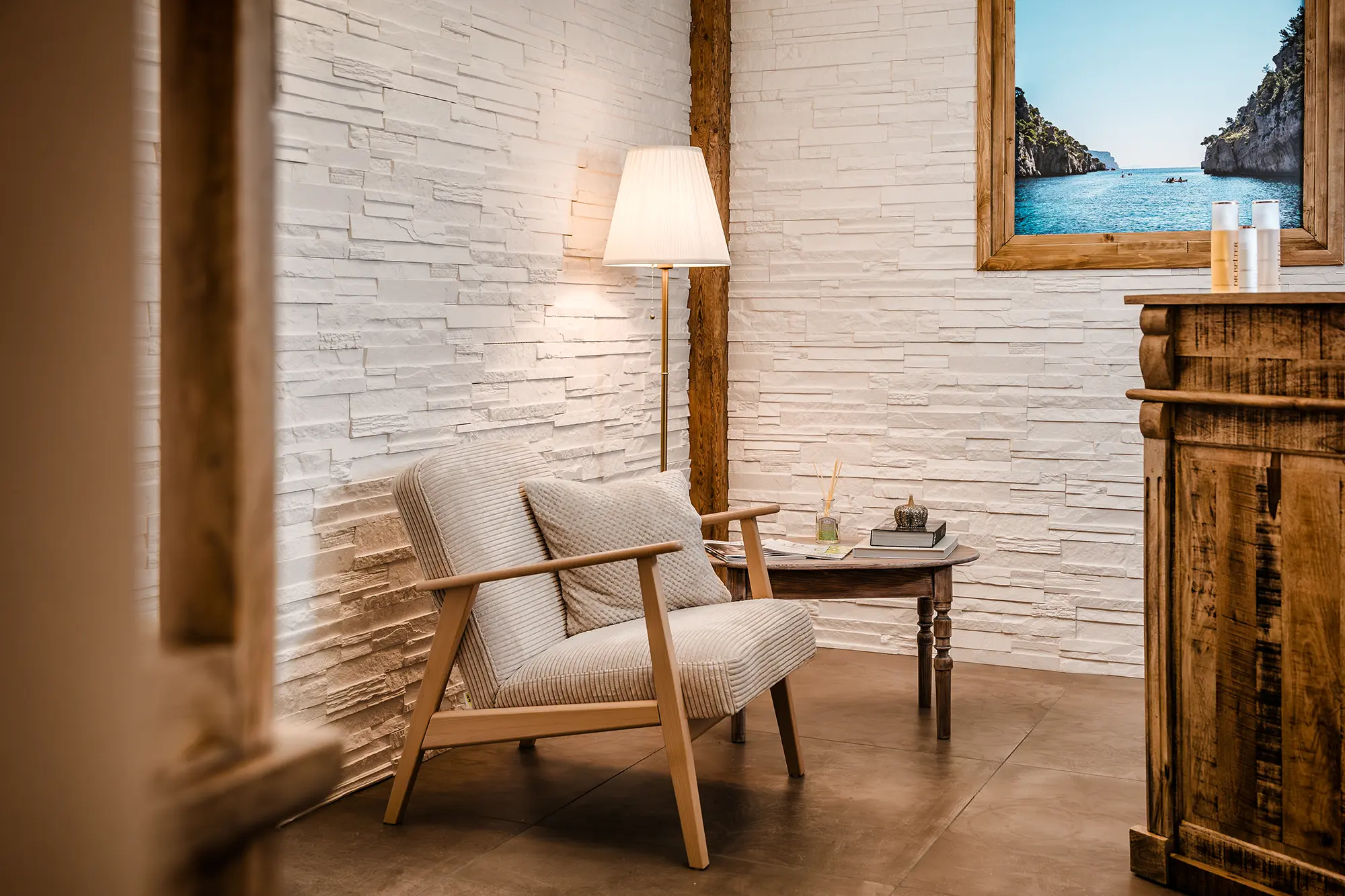 Wartebereich mit einem grauen Sessel, einem dunkelbraunen Beistelltisch aus Holz, einer großen Lampe mit weißem Schirm und einem großen Bild des Meeres an einer weißen Ziegelwand. Zur linken und rechten Seite ragen dunkelbraune Holzmöbel in das Bild.